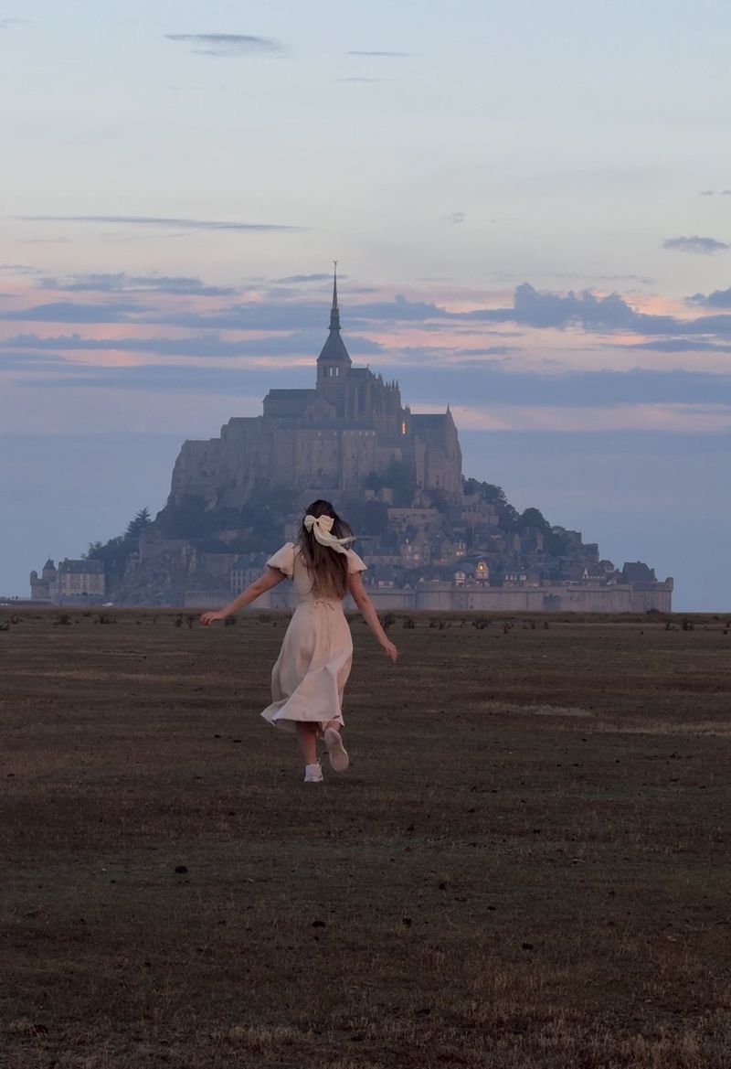 A woman in a light dress running across a dark field at dawn, with the silhouette of Mont-Saint-Michel behind her