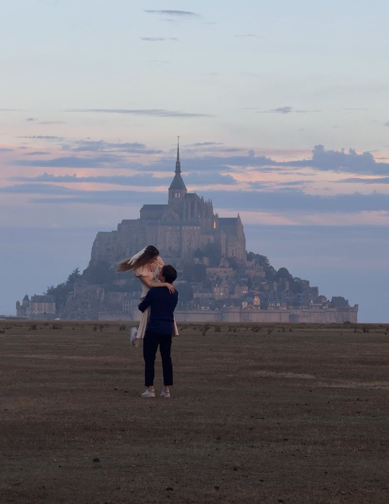 A man lifting a woman in a light dress in a field at dusk, with Mont-Saint-Michel behind them