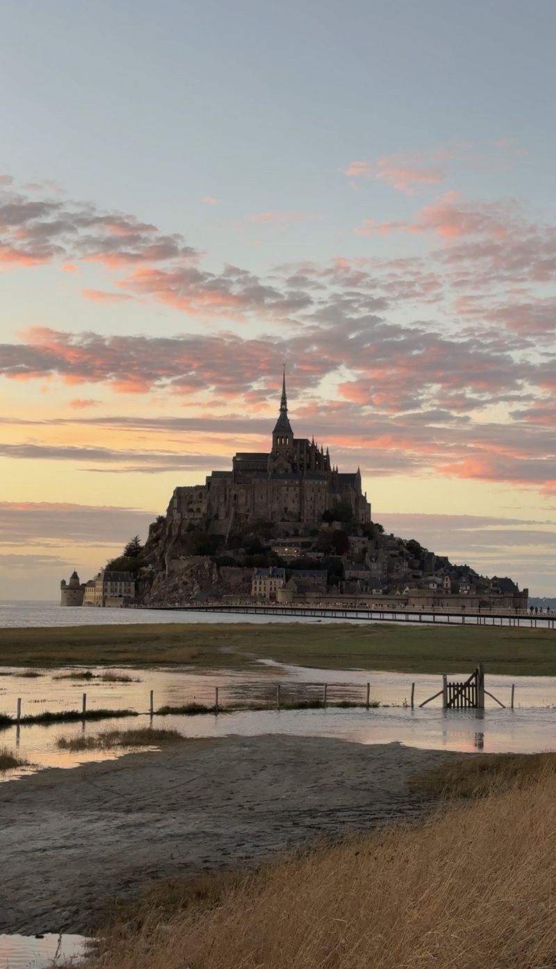 Mont-Saint-Michel at sunset under pink clouds, with tidal pools and a wooden fence in the foreground