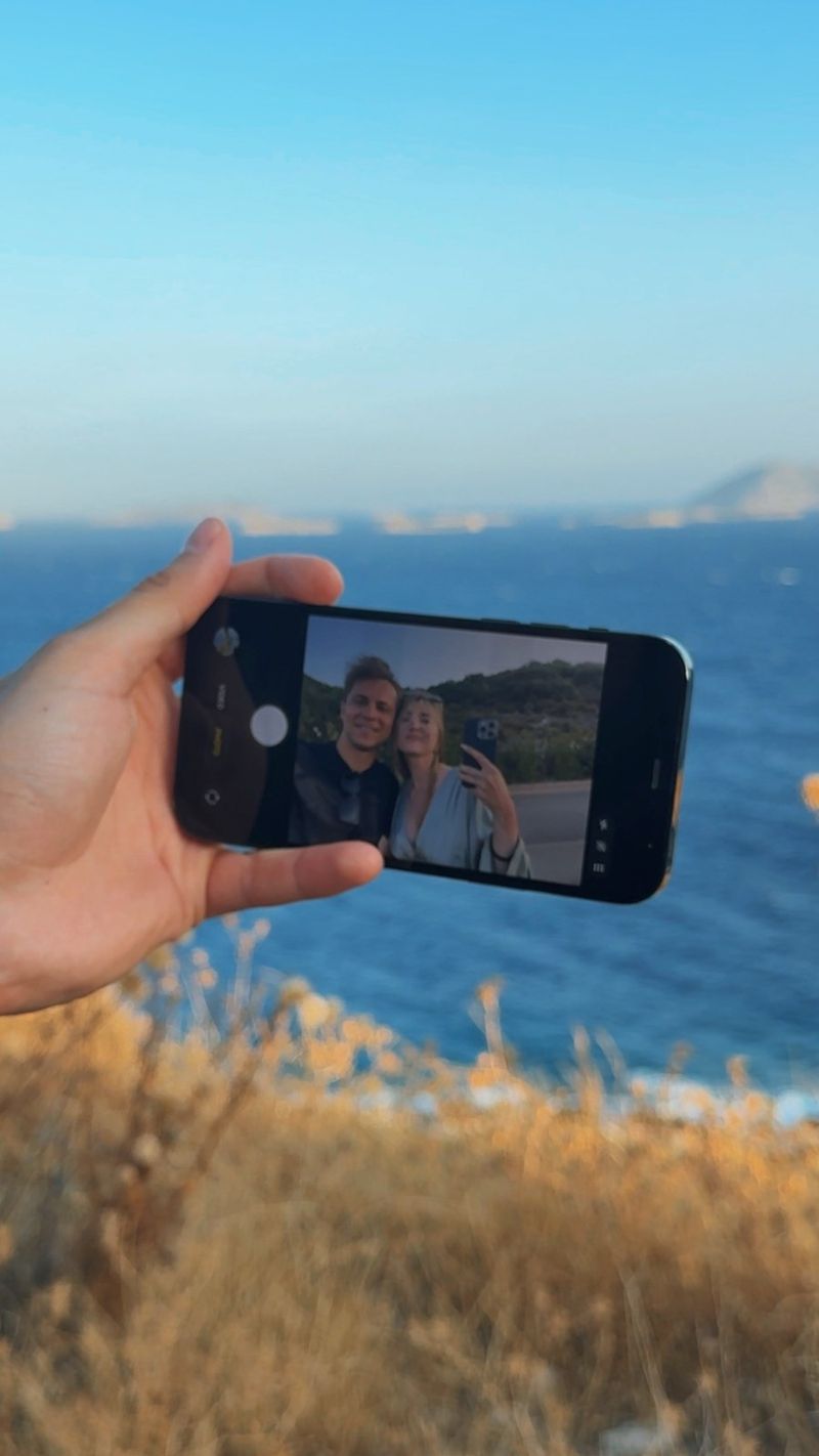 A hand holding up a phone against the blue sea, its screen showing a smiling couple taking a selfie