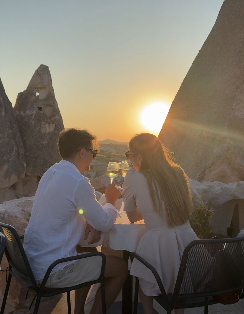 A couple toasting with glasses of white wine at a table among tall rock cones, the setting sun shining between them