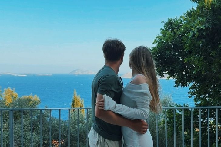 A couple with their arms around each other on a terrace, looking out over the blue sea and distant islands from Kaş