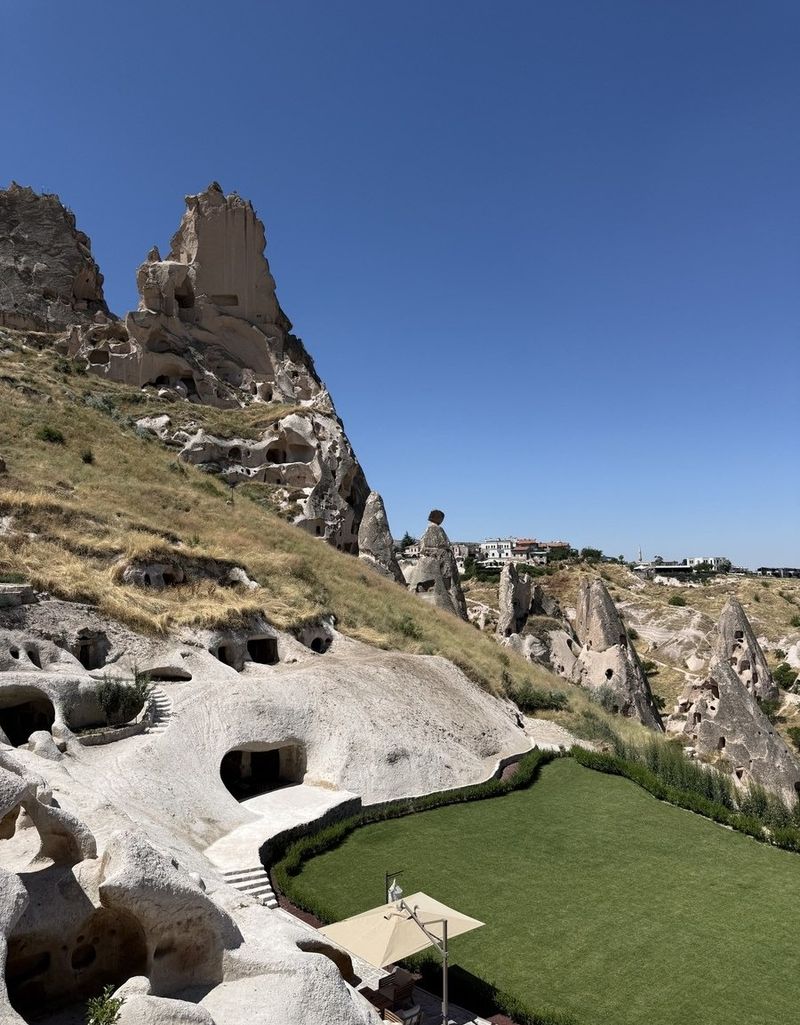 A cave hotel carved into a pale rock slope in Uçhisar, with a green lawn, a parasol and fairy chimneys under a deep blue sky