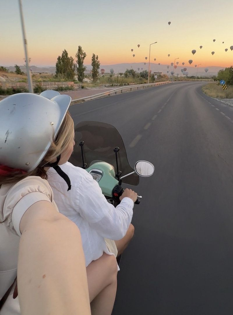 Over-the-shoulder view from the back of a scooter: a girl in a silver helmet with little ears riding along an empty road