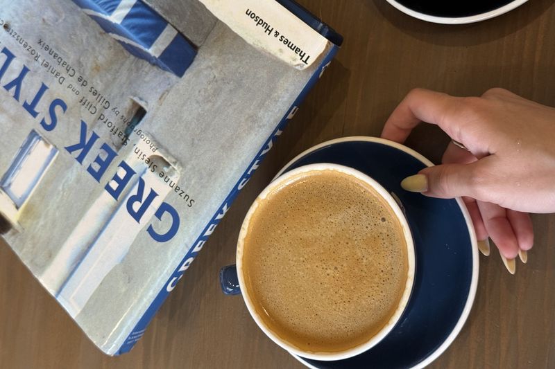 A hand holding a cup of cappuccino on a navy saucer beside a “Greek Style” coffee-table book on a wooden table