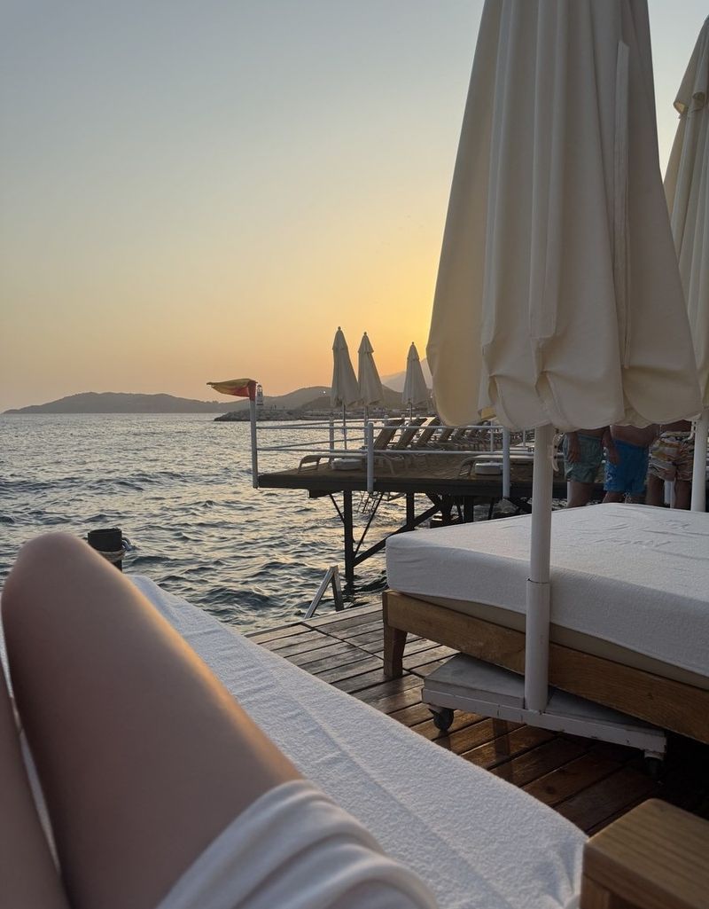 A wooden beach-club pier at sunset, with closed white parasols, sunbeds and the sea glowing gold beyond