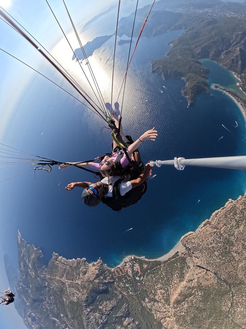 A tandem paraglider seen from above with a selfie stick, arms spread wide, high over the deep blue sea, green headlands and the curve of the Ölüdeniz coast
