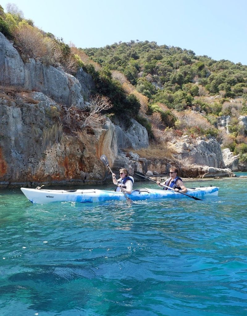 A couple paddling a white and blue tandem kayak past rocky limestone cliffs above turquoise water at Kekova