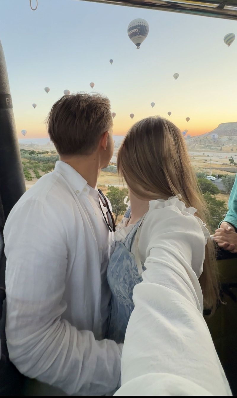 A couple in white shirts standing in a balloon basket at dawn, looking out at dozens of balloons over Cappadocia
